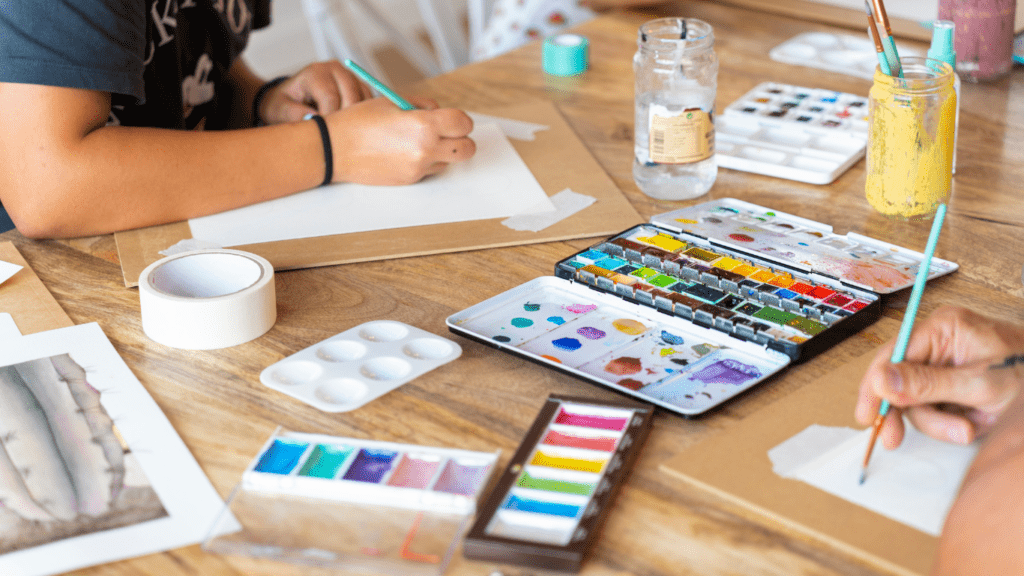 a group of people sitting at a table with watercolors and paintbrushes